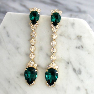 May include: A pair of long gold-tone drop earrings featuring a vertical chain of clear round crystals. Each earring is anchored by a pear-shaped emerald green crystal at the top and a larger matching pear-shaped emerald green crystal at the base.