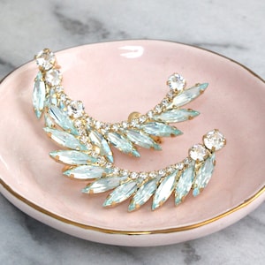 Puede incluir: Un par de pendientes de oro con detalles de cristal azul claro y transparente. Los pendientes tienen forma de media luna y están colocados en un plato rosa.