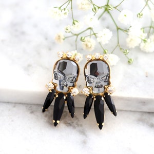 May include: A pair of gold tone earrings with black crystal accents. Each earring features a clear crystal skull with a gold tone setting.