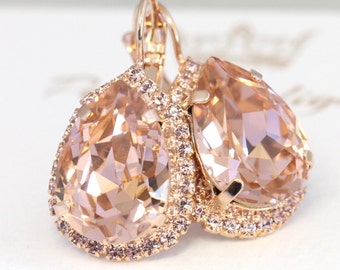 May include: Rose gold teardrop earrings with large peach-colored crystals. The crystals are surrounded by a halo of smaller, matching stones. The earrings have a lever back closure. The earrings are on a white surface.