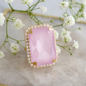 May include: A rectangular ring featuring a large, faceted light pink stone set in a gold-toned metal frame. The stone is surrounded by a border of small, clear, sparkling crystals and secured by four gold-toned prongs at the corners.