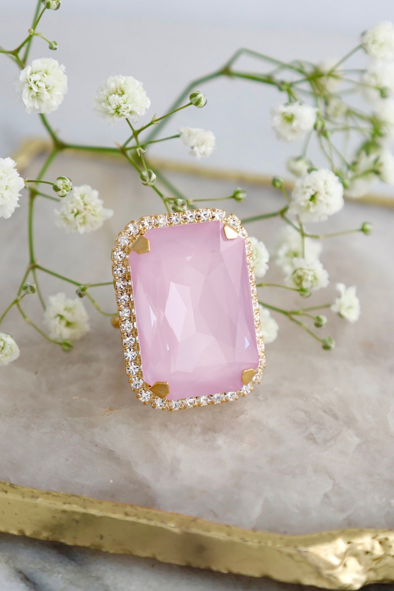 May include: A rectangular ring featuring a large, faceted light pink stone set in a gold-toned metal frame. The stone is surrounded by a border of small, clear, sparkling crystals and secured by four gold-toned prongs at the corners.