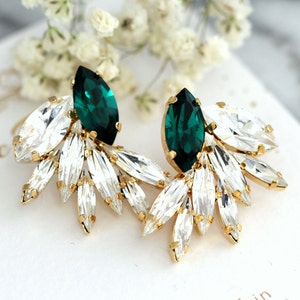 May include: A pair of gold-tone earrings featuring a central marquise-cut emerald green crystal surrounded by smaller clear marquise-cut crystals in a fan-like arrangement. The earrings are displayed on a white card with the text Ilona Rubin.