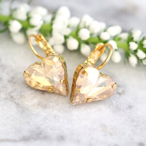 May include: A pair of gold-toned heart-shaped earrings with a faceted, champagne-colored crystal. The earrings are set on gold lever-back closures. The background features a sprig of white flowers and a marble surface.