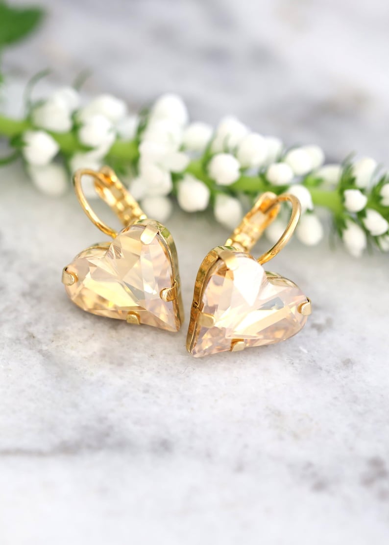 May include: A pair of gold-toned heart-shaped earrings with a faceted, champagne-colored crystal. The earrings are set on gold lever-back closures. The background features a sprig of white flowers and a marble surface.