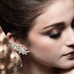 Op de afbeelding: Een close-up van een vrouwengezicht met dramatische oogmake-up. Haar ogen zijn gesloten en haar lippen zijn geschilderd in een metallic bruin. Ze draagt een glinsterende oorcuff.