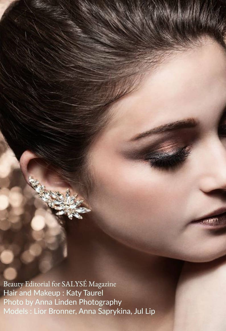 Op de afbeelding: Een close-up van een vrouwengezicht met dramatische oogmake-up. Haar ogen zijn gesloten en haar lippen zijn geschilderd in een metallic bruin. Ze draagt een glinsterende oorcuff.