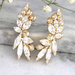 May include: A pair of gold earrings with clear and champagne-colored crystals arranged in a leaf-like design.