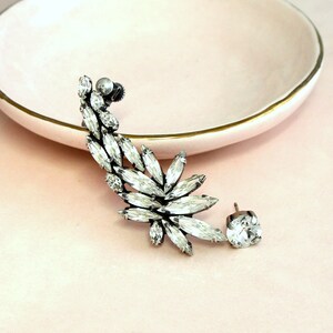 Op de afbeelding: Een zilveren oorcuff met een grote cluster van heldere strass steentjes. De cuff is ontworpen om om het oor te wikkelen en heeft een enkele strass steentje.