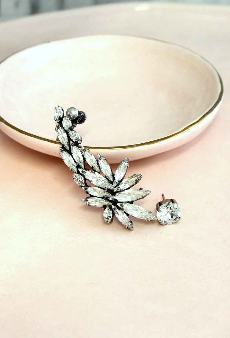 Op de afbeelding: Een zilveren oorcuff met een grote cluster van heldere strass steentjes. De cuff is ontworpen om om het oor te wikkelen en heeft een enkele strass steentje.