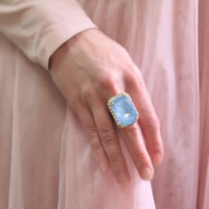 May include: A large, light blue, rectangular ring with a gold setting and small clear stones around the edge. The ring is on a finger wearing a pink dress.