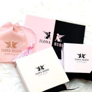 May include: An assortment of Ilona Rubin petite delights packaging, including a pink drawstring bag, pink and black cards, and white boxes. Each item features the brand's logo of winged figures. The items are arranged on a white, fluffy surface.