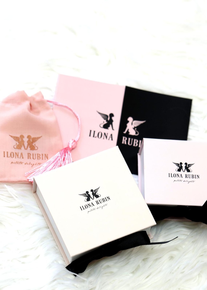 May include: An assortment of Ilona Rubin petite delights packaging, including a pink drawstring bag, pink and black cards, and white boxes. Each item features the brand's logo of winged figures. The items are arranged on a white, fluffy surface.