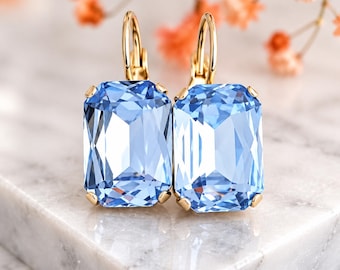 May include: A pair of gold leverback earrings featuring large, rectangular, faceted light blue crystals. The earrings are positioned upright, showcasing the clear, geometric cut of the stones and the polished gold metal settings that secure each crystal with four small prongs.