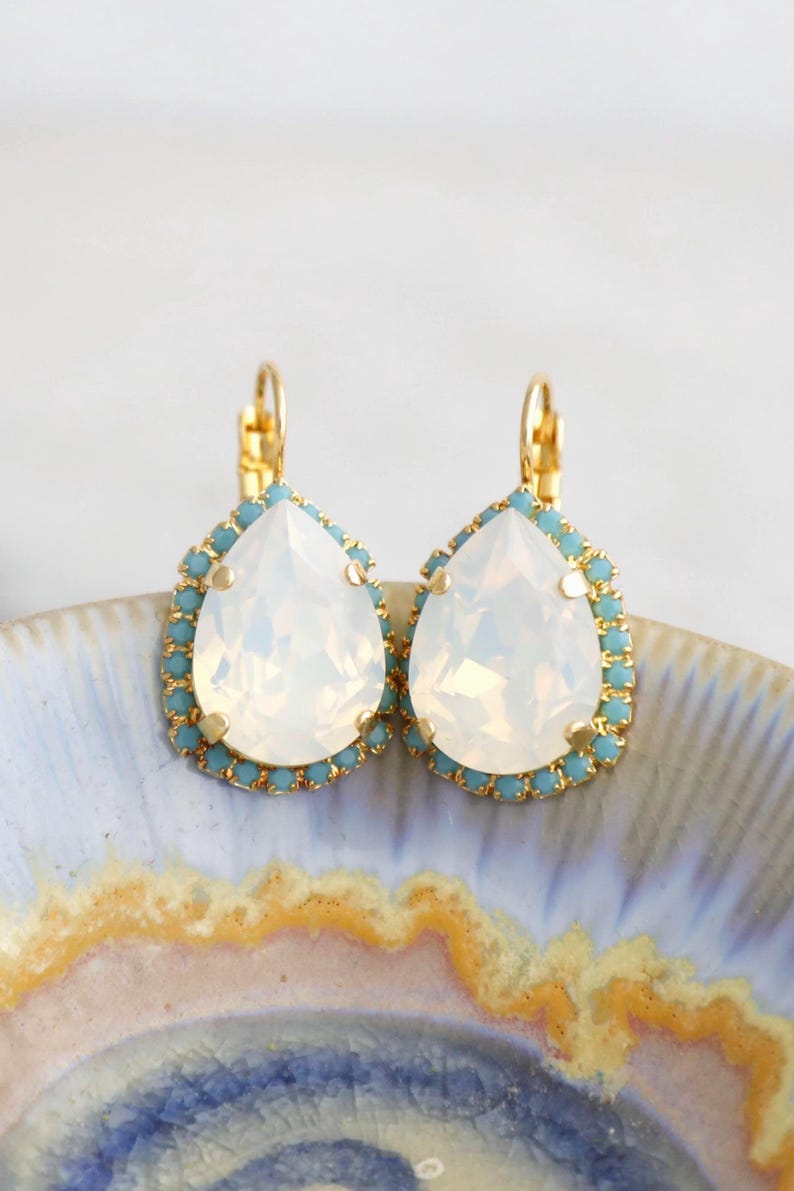 May include: A pair of gold teardrop earrings with a white opal stone and a light blue crystal border.