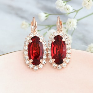 May include: A pair of oval-shaped earrings featuring a large, deep red faceted center stone surrounded by a halo of small, clear white crystals. The stones are set in a rose gold-toned metal frame with a secure lever-back closure mechanism.