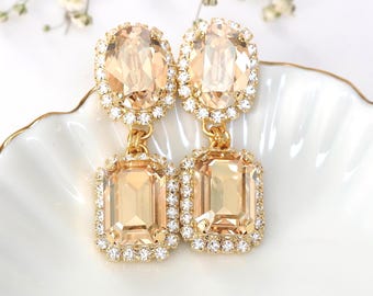 May include: A pair of gold-toned drop earrings with peach-colored, rectangular and oval-shaped crystals, surrounded by smaller clear crystals. The earrings are embellished with Swarovski crystals.