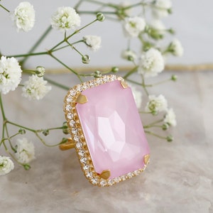 May include: A gold-toned ring featuring a large, rectangular, faceted light pink stone set in a frame of small, clear, sparkling crystals. The stone is secured by four gold-toned prongs at the corners, creating a bold and elegant jewelry piece.