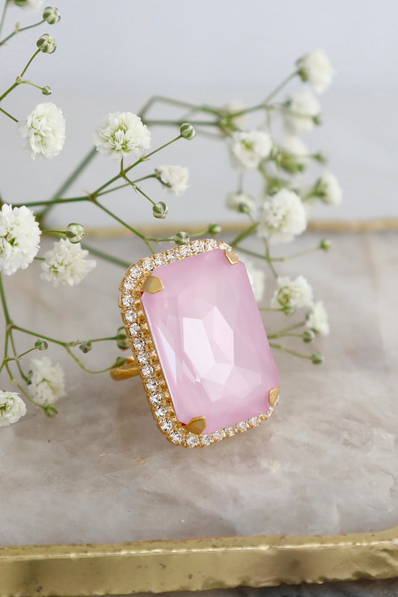May include: A gold-toned ring featuring a large, rectangular, faceted light pink stone set in a frame of small, clear, sparkling crystals. The stone is secured by four gold-toned prongs at the corners, creating a bold and elegant jewelry piece.