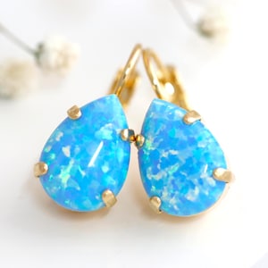 May include: A pair of teardrop-shaped blue opal earrings set in gold-toned metal prongs. Each earring features a vibrant, iridescent blue stone with a speckled pattern, secured by a gold-toned lever back mechanism. The brand name Ilona Rubin is printed at the bottom.