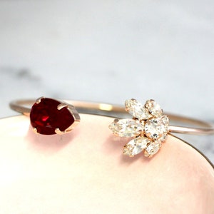 May include: A gold bangle bracelet with a red gemstone on one end and a cluster of clear rhinestones on the other end.
