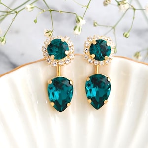 May include: Emerald green teardrop earrings with gold-tone settings. Each earring features a round, crystal-studded top and a large, pear-shaped crystal pendant. The earrings are displayed on a white surface with floral accents. Text: embellished with Swarovski crystals.