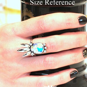 Puede incluir: Un anillo de plata con un dise&ntilde;o de cristal transparente y un camafeo colorido. El anillo est&aacute; en un dedo y el texto "Size Reference" es visible en el fondo.