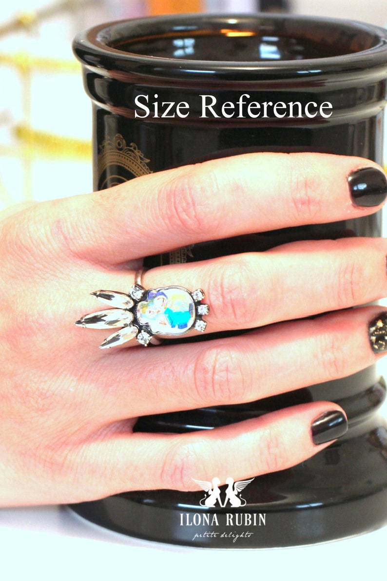 Puede incluir: Un anillo de plata con un dise&ntilde;o de cristal transparente y un camafeo colorido. El anillo est&aacute; en un dedo y el texto "Size Reference" es visible en el fondo.