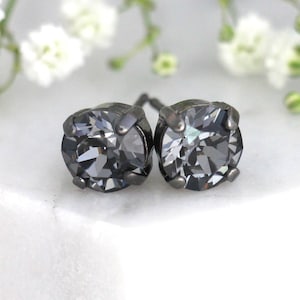 Puede incluir: Un par de pendientes de botón redondos con cristales facetados de color gris oscuro en un engaste de metal oscuro. Las piedras circulares tienen una superficie reflectante con múltiples facetas que captan la luz, creando un aspecto de carbón metálico.