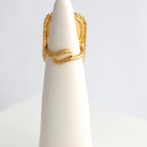 May include: A gold ring featuring a rectangular face with a textured border and a smooth, polished finish. The band is open and adjustable, characterized by a delicate, curved, wave-like design that wraps around the finger. The metal has a bright, reflective yellow gold tone throughout the entire piece.