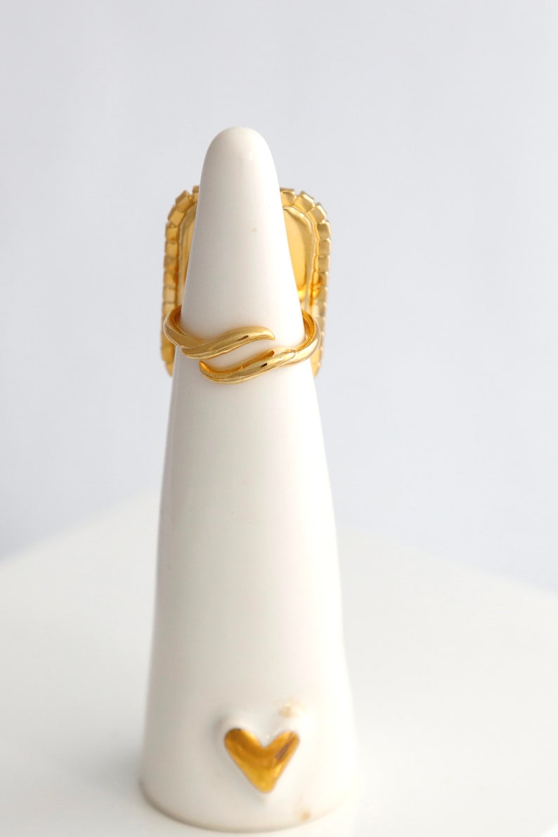 May include: A gold ring featuring a rectangular face with a textured border and a smooth, polished finish. The band is open and adjustable, characterized by a delicate, curved, wave-like design that wraps around the finger. The metal has a bright, reflective yellow gold tone throughout the entire piece.
