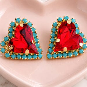 May include: A pair of heart-shaped earrings featuring large, faceted red crystals set in gold-tone metal. Each earring is bordered by a row of small, round, bright turquoise beads. The text ILONA RUBIN petite delights Atelier-made jewelry is visible.