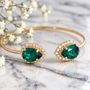 May include: A gold-toned open cuff bracelet featuring two pear-shaped emerald green crystals, each surrounded by a halo of small clear stones. The piece is marked with the text ILONA RUBIN Atelier-made jewelry at the bottom.