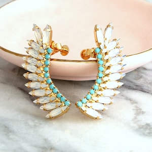 Puede incluir: Un par de puños de oreja dorados. Cada puño presenta un diseño curvo con acentos blancos en forma de cristal y pequeñas cuentas de color turquesa. Los pendientes se exhiben en un plato rosa, sobre una superficie de mármol.