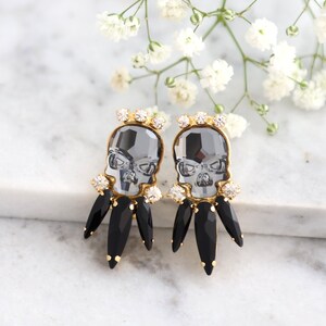 May include: A pair of gold tone earrings featuring a black crystal skull with clear crystal accents and black crystal teardrop dangles.