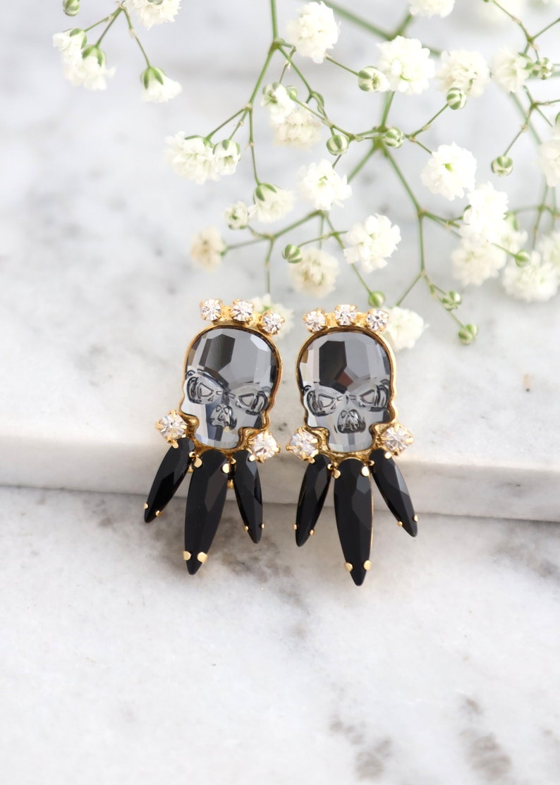 May include: A pair of gold tone earrings featuring a black crystal skull with clear crystal accents and black crystal teardrop dangles.