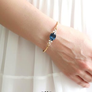 May include: A gold-toned bracelet with a large, oval, dark blue gemstone as the focal point. The bracelet is embellished with smaller, clear crystals. The image includes the text "ILONA RUBIN petite delights" and "embellished with Swarovski crystals".