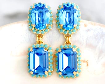 Puede incluir: Pendientes colgantes con cristales azules claros ovalados y rectangulares, rodeados de pequeñas piedras turquesas y montados en metal dorado. Están adornados con cristales Swarovski. El diseño es de doble caída.