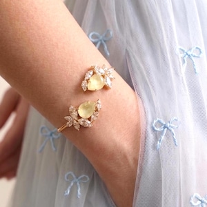 Op de afbeelding: Een goudkleurige manchetarmband met twee peervormige gele edelstenen, elk omringd door heldere kristallen. De armband wordt om een pols gedragen, tegen een lichtblauwe achtergrond. De tekst "ILONA RUBIN" en "Atelier-made jewelry" is zichtbaar.