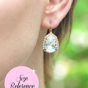 May include: Gold teardrop earrings with a large clear crystal surrounded by smaller crystals. The earrings are dangling from the earlobe.