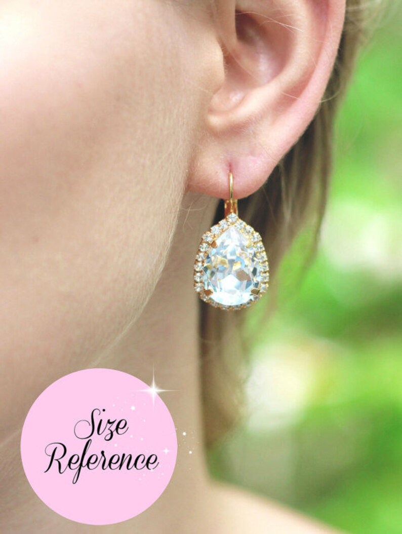 May include: Gold teardrop earrings with a large clear crystal surrounded by smaller crystals. The earrings are dangling from the earlobe.