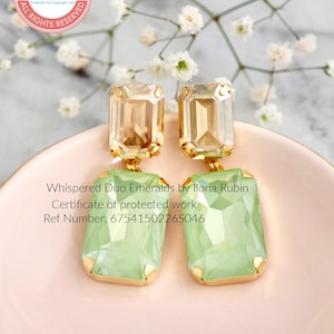 Może przedstawiać: Prostokątne kolczyki w kolorze jasnozielonym, z oprawami w kolorze złota. Kolczyki są oznaczone jako "Whispered Duo Emeralds by Ilona Rubin" i prezentowane na r&oacute;żowej powierzchni.
