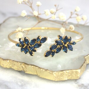 May include: A gold bangle bracelet with two clusters of dark blue marquise-shaped rhinestones. The bracelet is open on one side and has a delicate, feminine design.