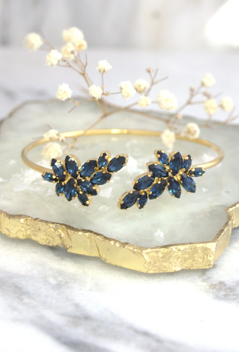 May include: A gold bangle bracelet with two clusters of dark blue marquise-shaped rhinestones. The bracelet is open on one side and has a delicate, feminine design.