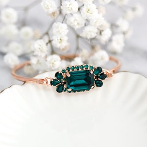 May include: Rose gold bangle bracelet with a large emerald-cut, emerald-green gemstone center and smaller surrounding stones. The bracelet is displayed on a white, scalloped dish with white flowers in the background.