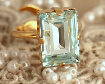 May include: A gold ring with a large, light blue, emerald-cut gemstone. The ring is set on a white lace background.