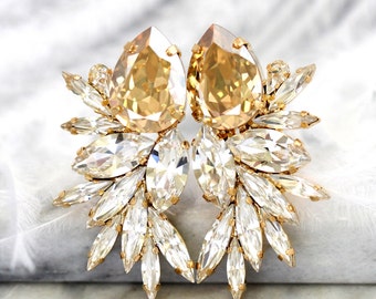 May include: A pair of gold earrings with a large teardrop-shaped crystal in the center of each earring. The crystals are surrounded by smaller, clear crystals that are arranged in a fan shape.