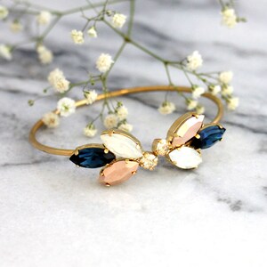 May include: Gold bangle bracelet with a cluster of blue, white, and pink crystal stones in a leaf shape.