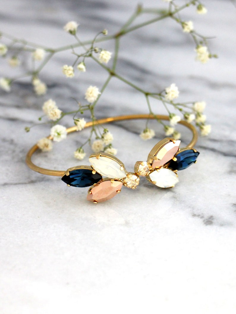 May include: Gold bangle bracelet with a cluster of blue, white, and pink crystal stones in a leaf shape.