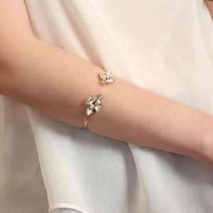 May include: A gold cuff bracelet with a delicate design featuring clear crystal accents. The bracelet is open on one side and has a small cluster of crystals on the other side.